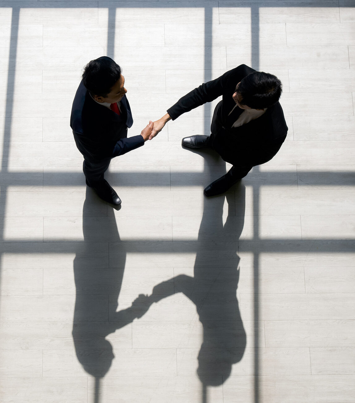 Business partners shaking hands in the modern office. Standing near the window while the sunlight shines on them. There is a shadow on the wooden floor