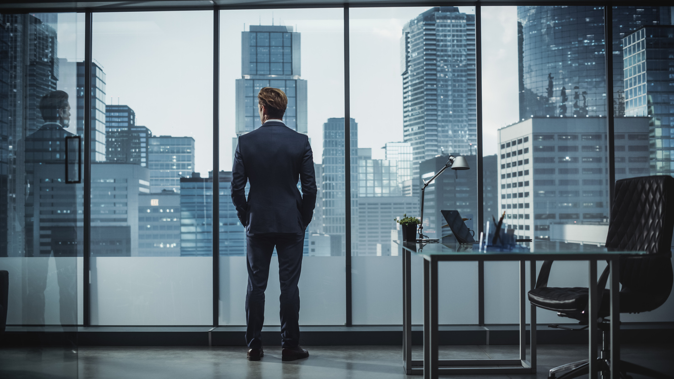 Successful Young Businessman in a Perfect Tailored Suit Standing in His Modern Office Looking out of the Window on Big City with Skyscrapers. Successful Finance Manager Planning Project Strategy.